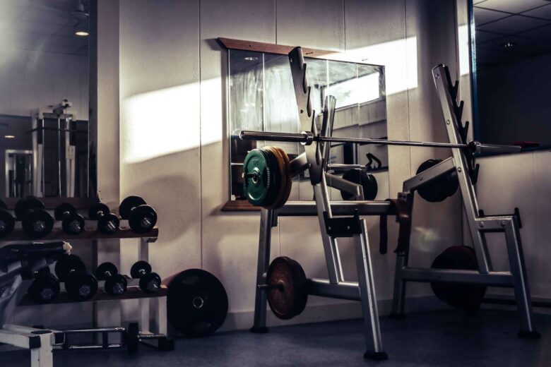 A gym with a loaded barbell on a bench press rack, dumbbells on a nearby rack, weight plates on the floor, and a large wall mirror reflecting light. The room has a calm, empty atmosphere.