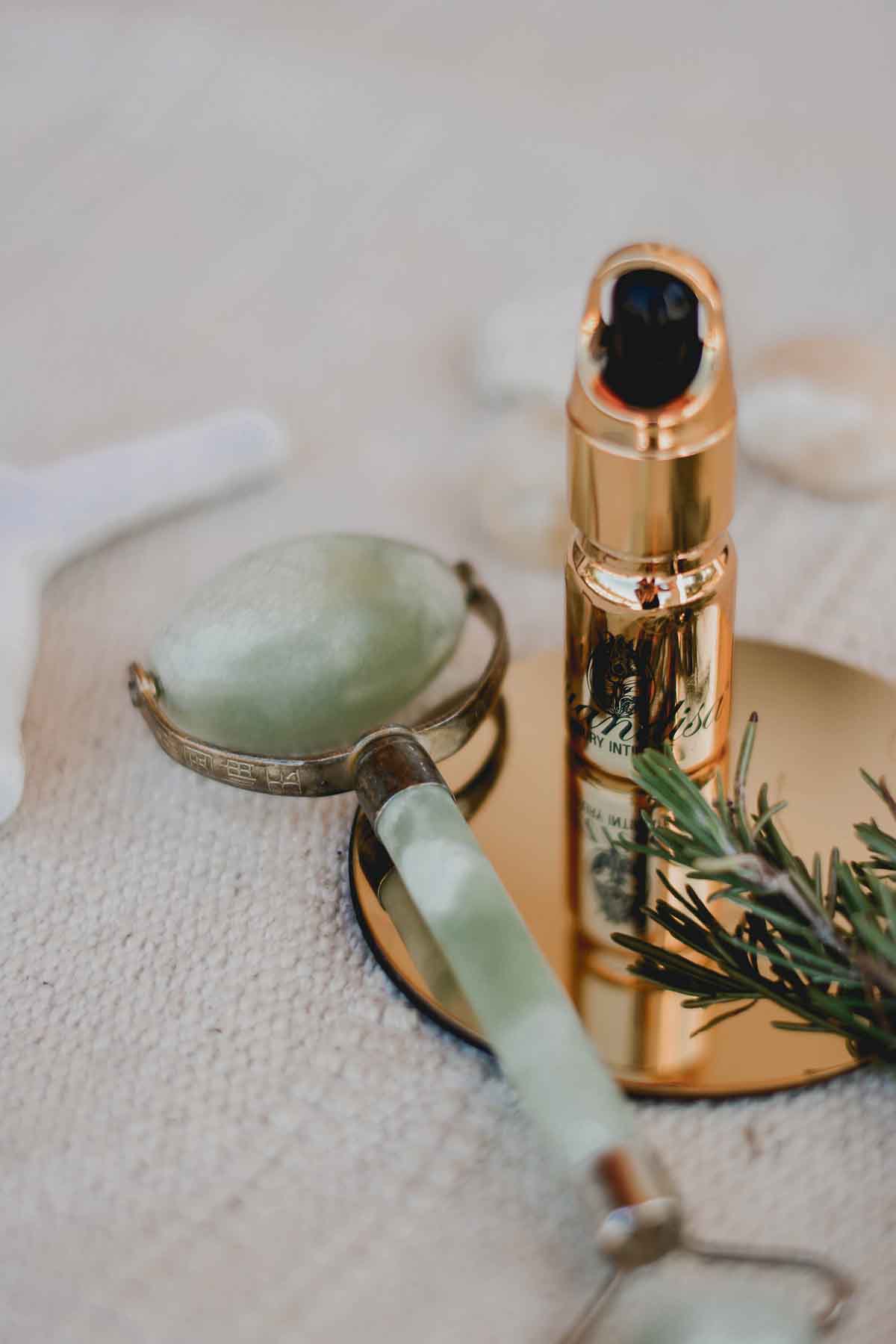 A jade facial roller and a gold skincare serum bottle rest on a round gold tray, with a sprig of rosemary nearby, all placed on a textured beige surface.