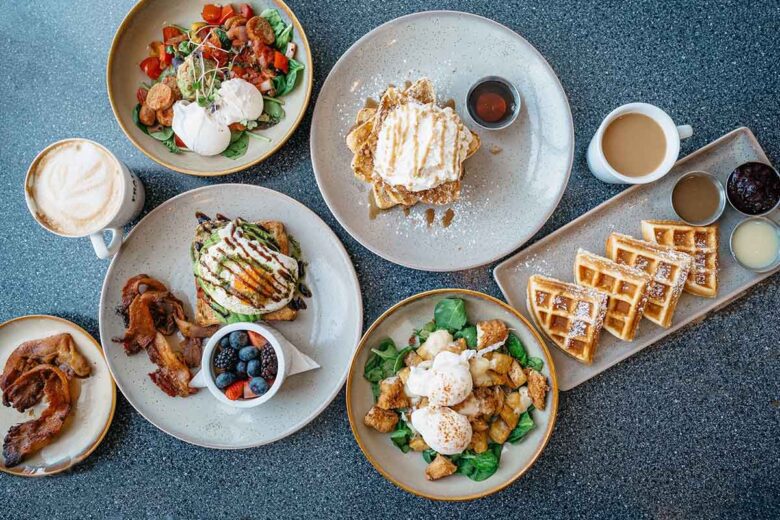 A table set with various breakfast dishes, including waffles, pancakes, eggs with toast, a bowl of fruit, bacon, and cups of coffee, all arranged on gray speckled tabletop.