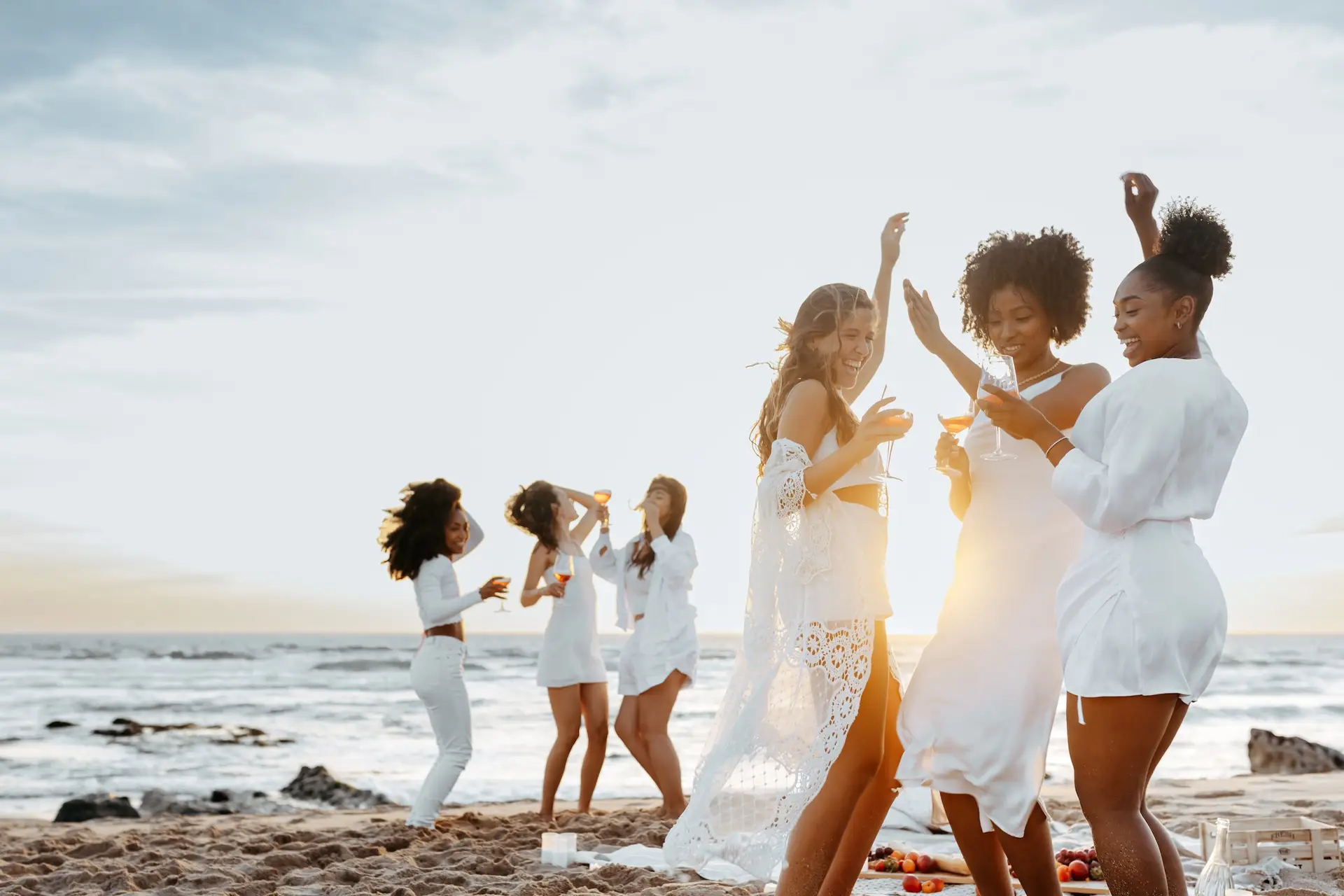 A group of women dressed in white dance, laugh, and enjoy drinks together on a sandy beach at sunset, with the ocean and a bright sky in the background.