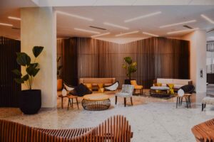 A modern, stylish lobby with neutral and yellow furniture, wood slat walls, potted plants, and terrazzo flooring, creating a warm and inviting atmosphere.
