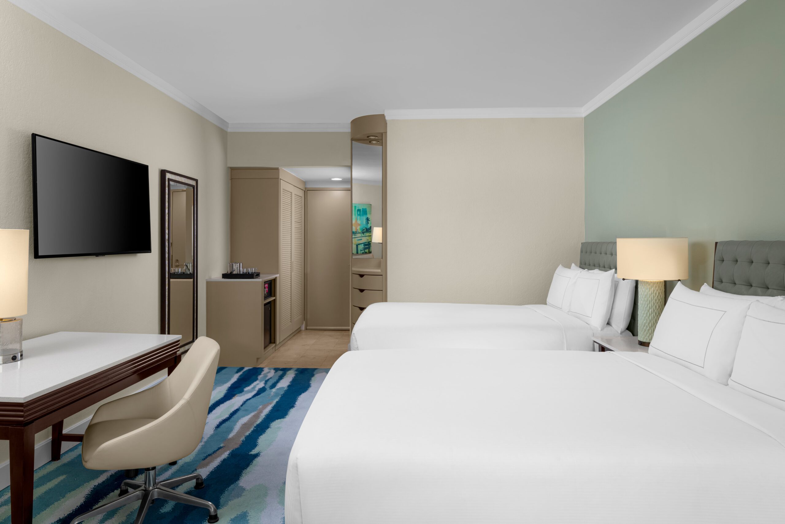 Modern hotel room with two double beds, a desk and chair, large wall-mounted TV, dresser with a mirror, mini-fridge, lamps, and a closet. The room features light walls and a blue patterned carpet.