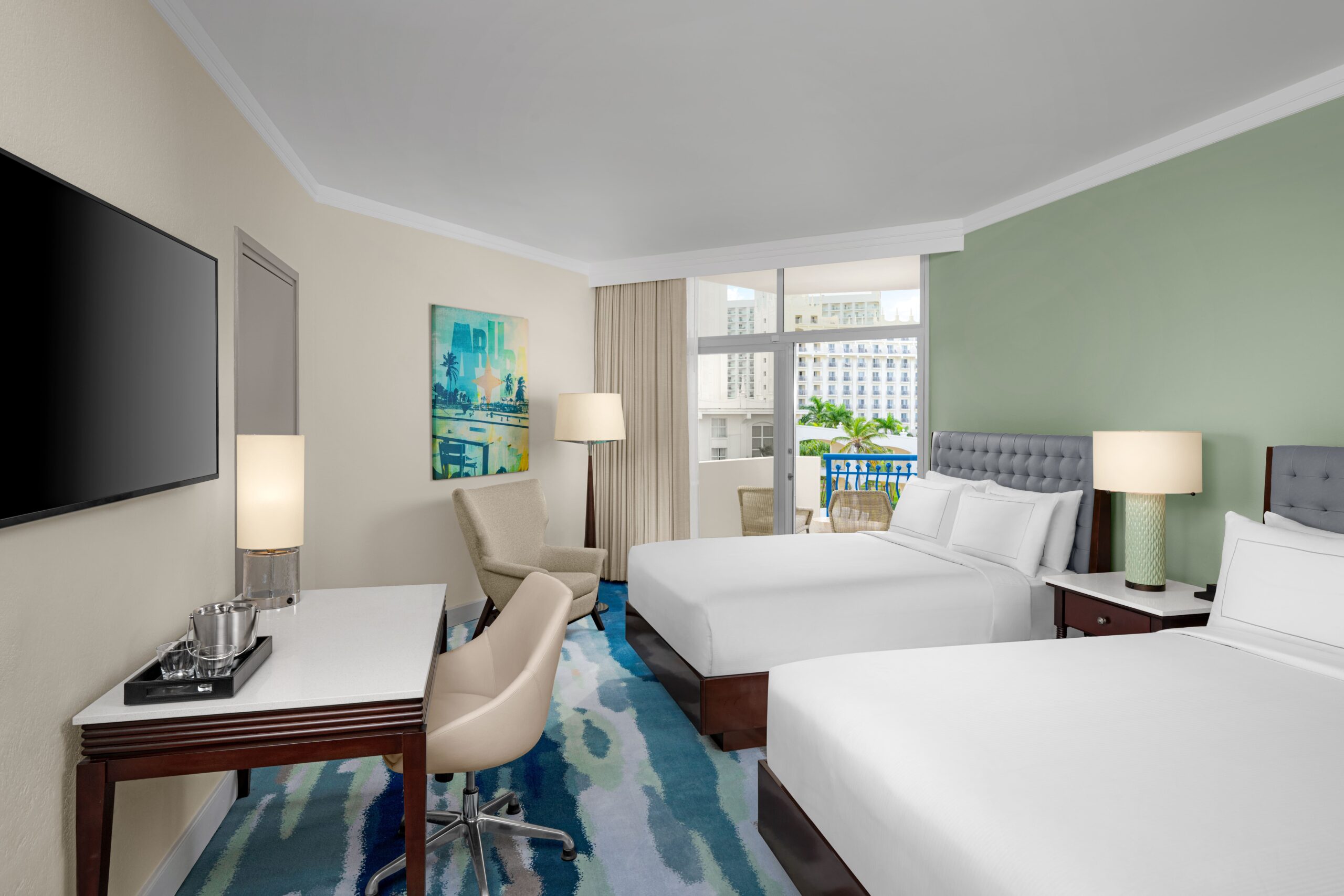 A modern hotel room with two double beds, a desk and chair, a flat-screen TV, lamps, an armchair, colorful artwork, and large window doors opening to a balcony with city views.