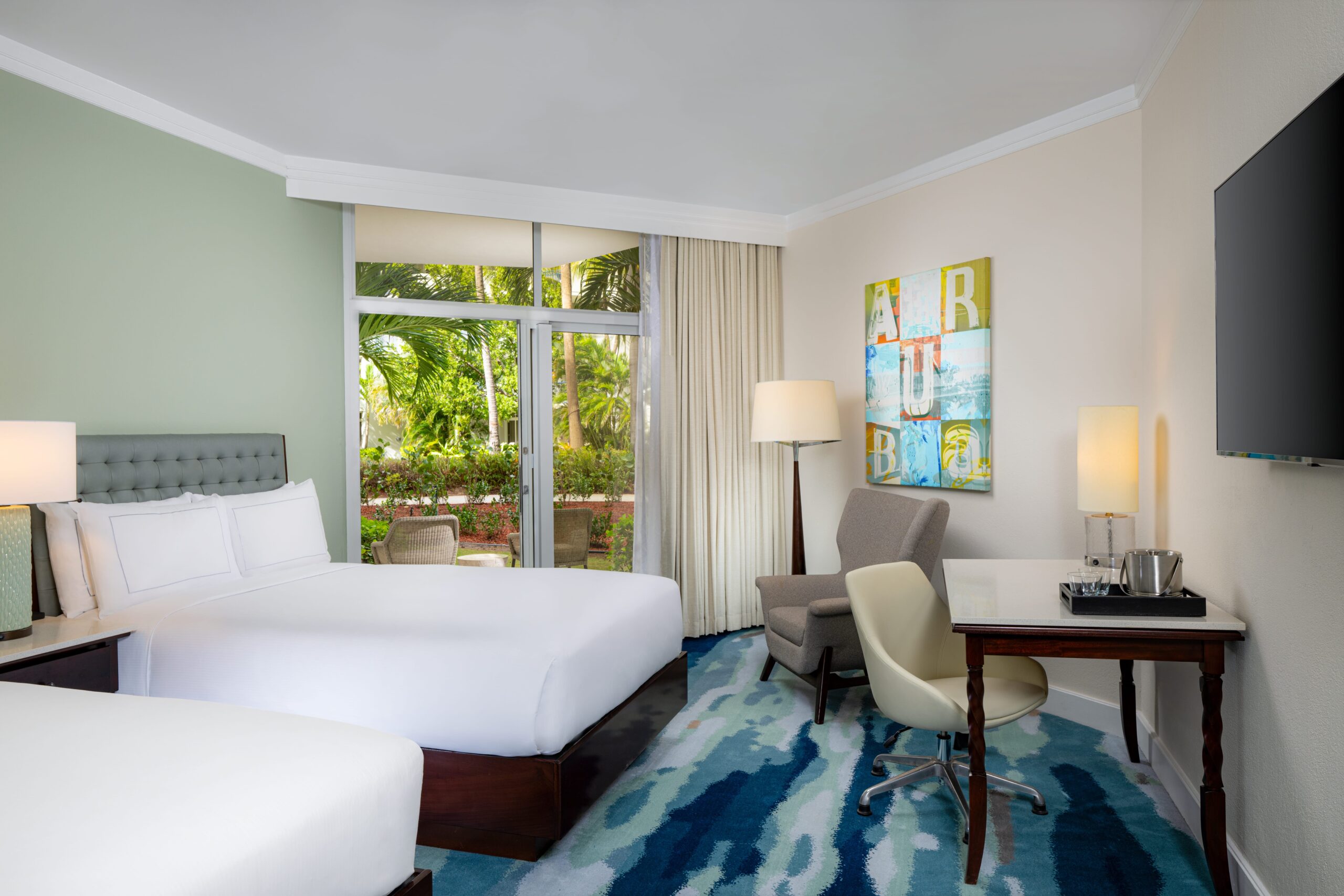 Bright hotel room with two double beds, a desk and chair, an armchair with a floor lamp, a wall-mounted TV, and large windows with a view of lush greenery outside. Coastal-themed decor is visible on the wall.