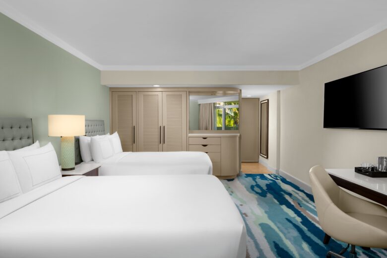 A modern hotel room with two large beds, a nightstand with a lamp, a flat-screen TV on the wall, a desk with a chair, and a dresser near a mirrored closet; a window shows greenery outside.