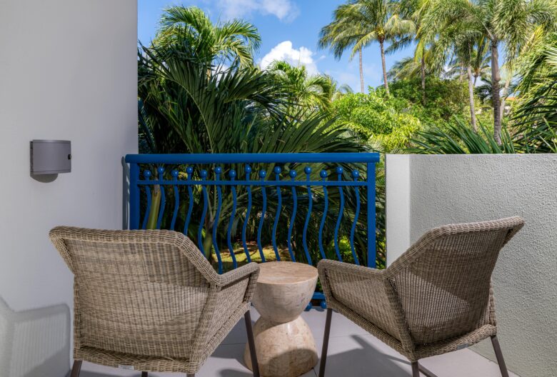 Two wicker chairs and a small round table are set on a balcony with a blue railing, overlooking lush green palm trees under a sunny, partly cloudy sky.