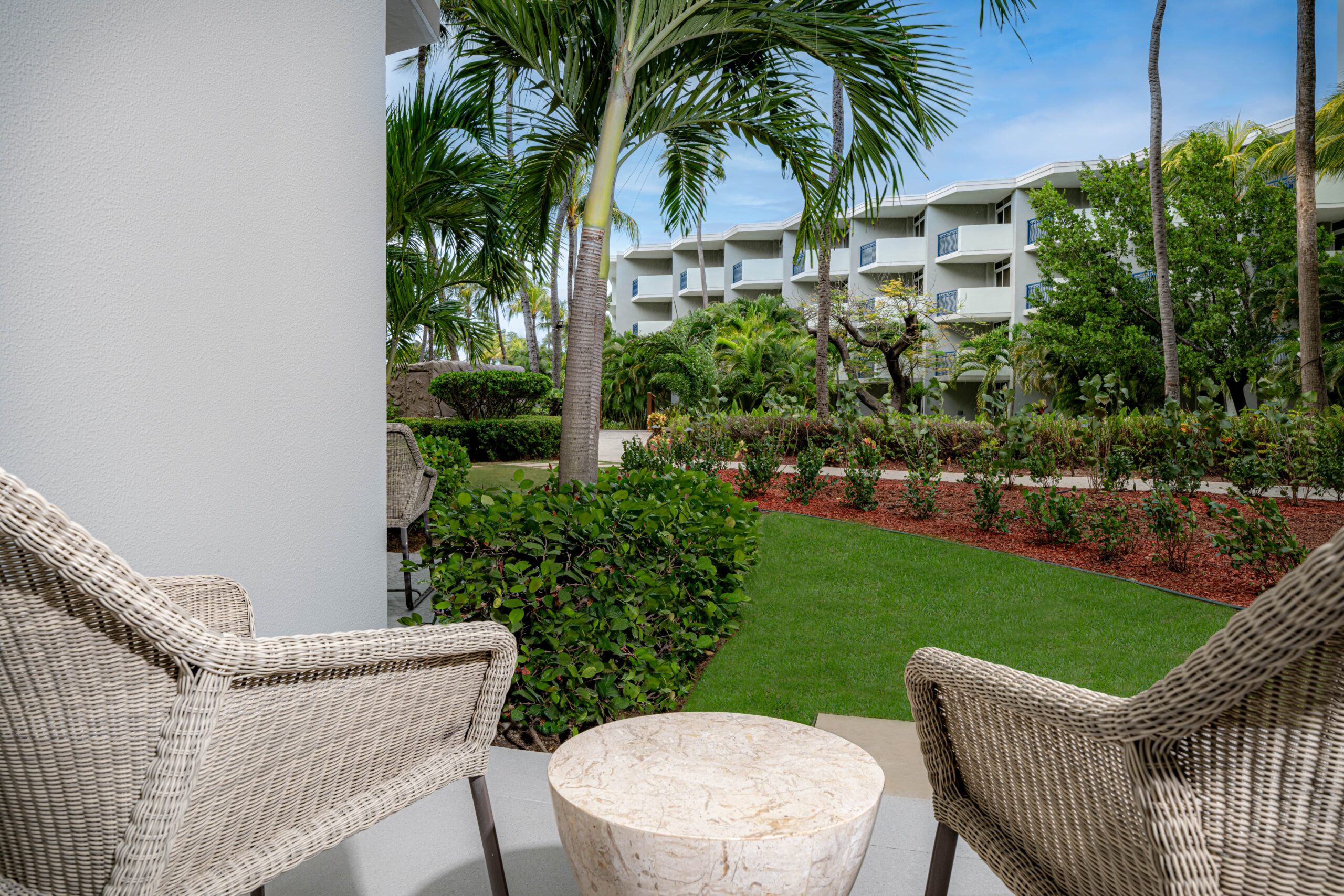 Two wicker chairs and a small round table sit on a patio overlooking a garden with palm trees and green grass, with a modern white apartment building visible in the background.