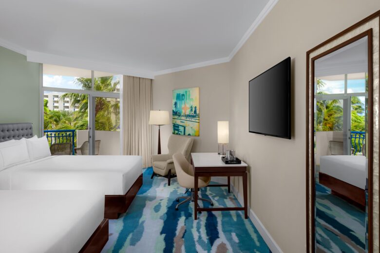 Modern hotel room with two double beds, a desk and chair, a large wall-mounted TV, a floor lamp, and a window overlooking palm trees. Room features colorful carpet and contemporary artwork.
