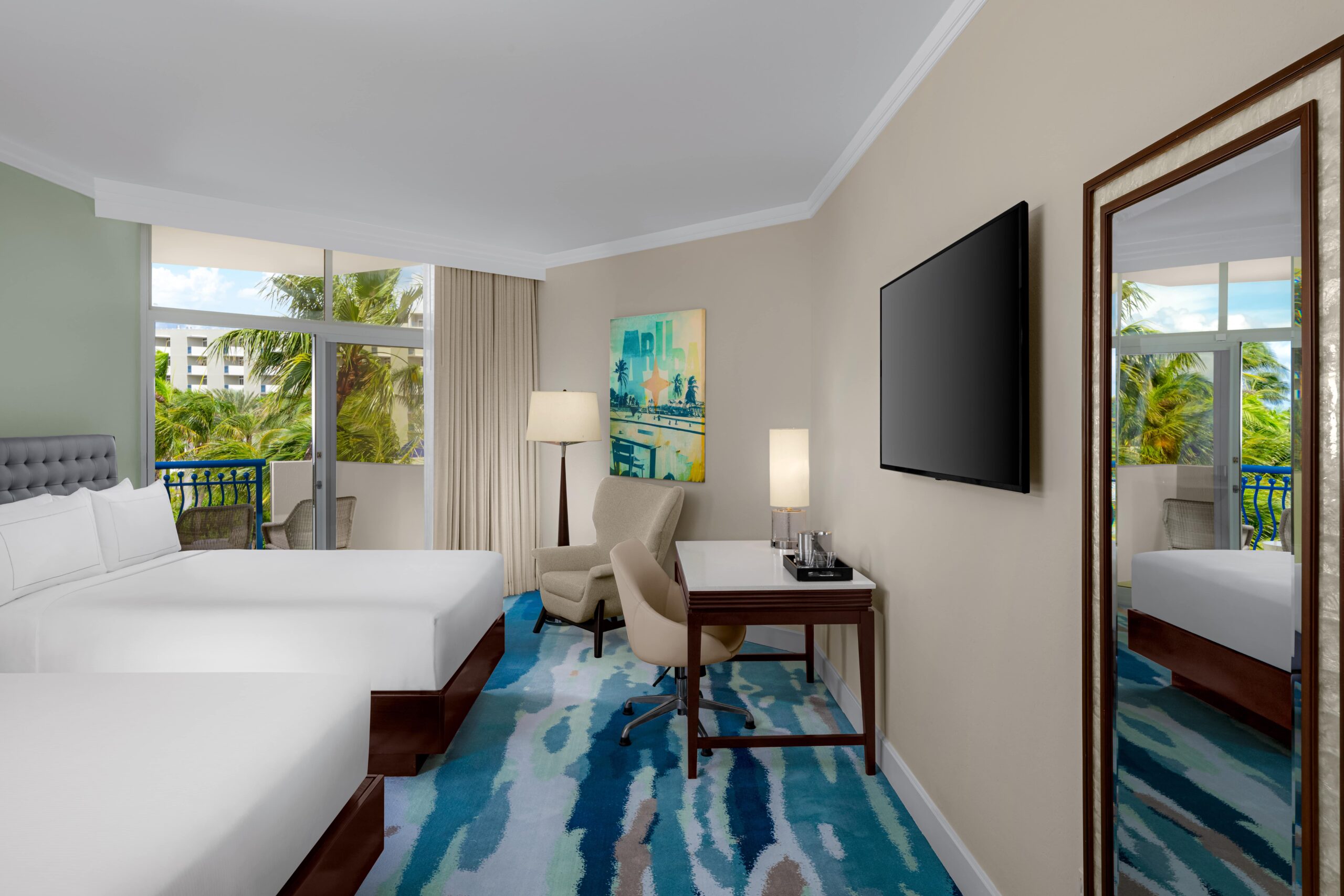 Modern hotel room with two double beds, a desk and chair, a large wall-mounted TV, a floor lamp, and a window overlooking palm trees. Room features colorful carpet and contemporary artwork.