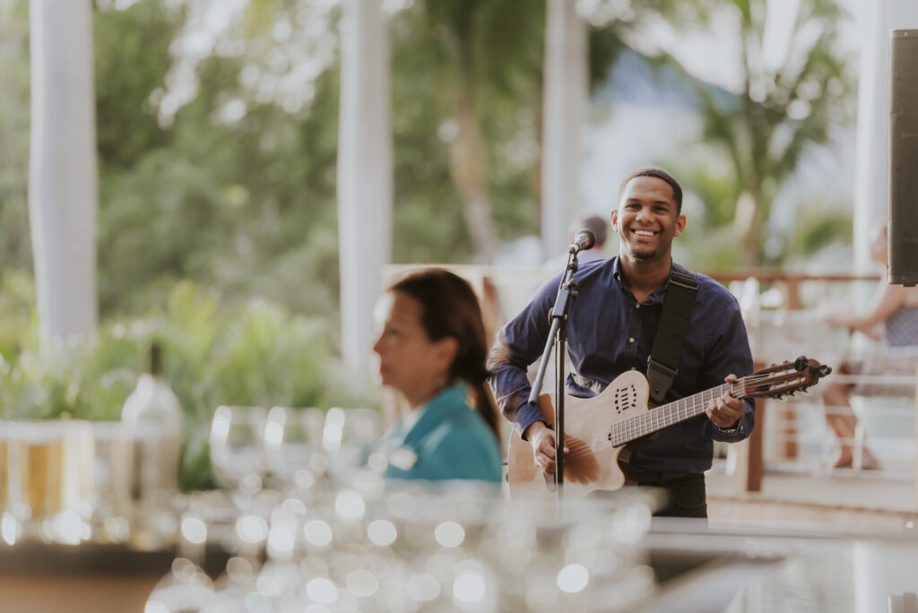 A smiling man plays guitar and sings into a microphone outdoors, with blurred glasses and a woman in the foreground, and green trees in the background.