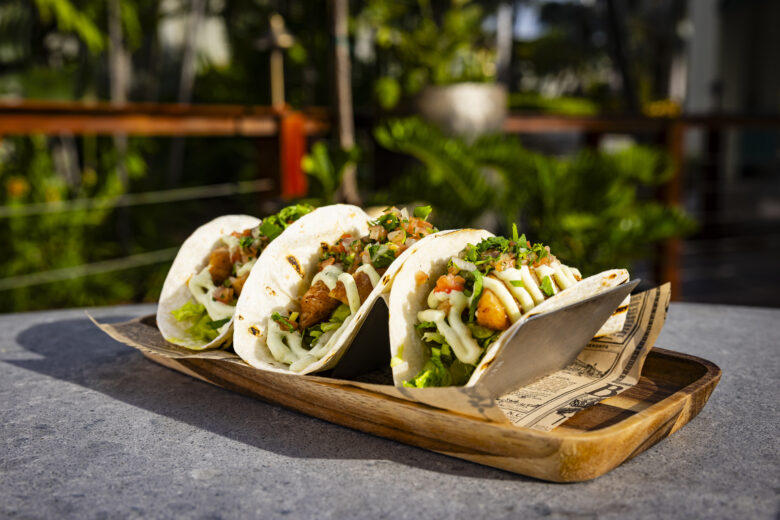 Three soft shell tacos filled with lettuce, diced tomatoes, grilled fish, chopped cilantro, and drizzled with a creamy sauce, served on a wooden tray outdoors with greenery in the background.