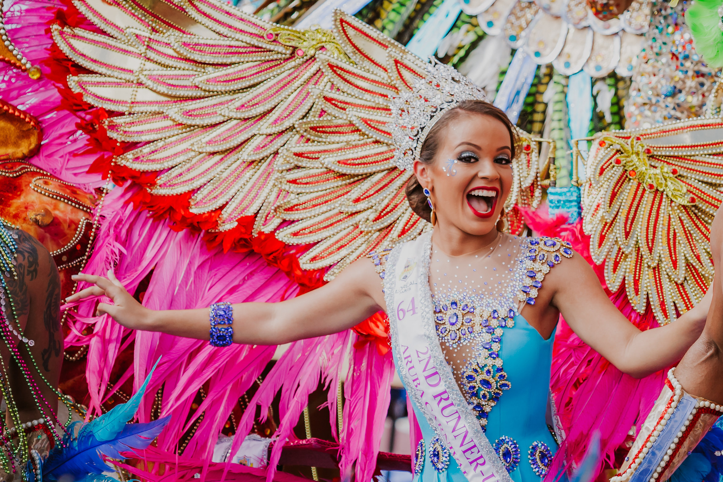 A woman in a vibrant blue costume with jeweled details, a crown, colorful feathered wings, and a sash reading "2nd Runner Up" smiles and poses joyfully at a lively carnival or parade.