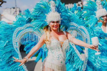 A woman in a sparkling costume with blue feathered wings and a beaded headdress smiles and poses outdoors, holding a white cup, during a festive parade or carnival.