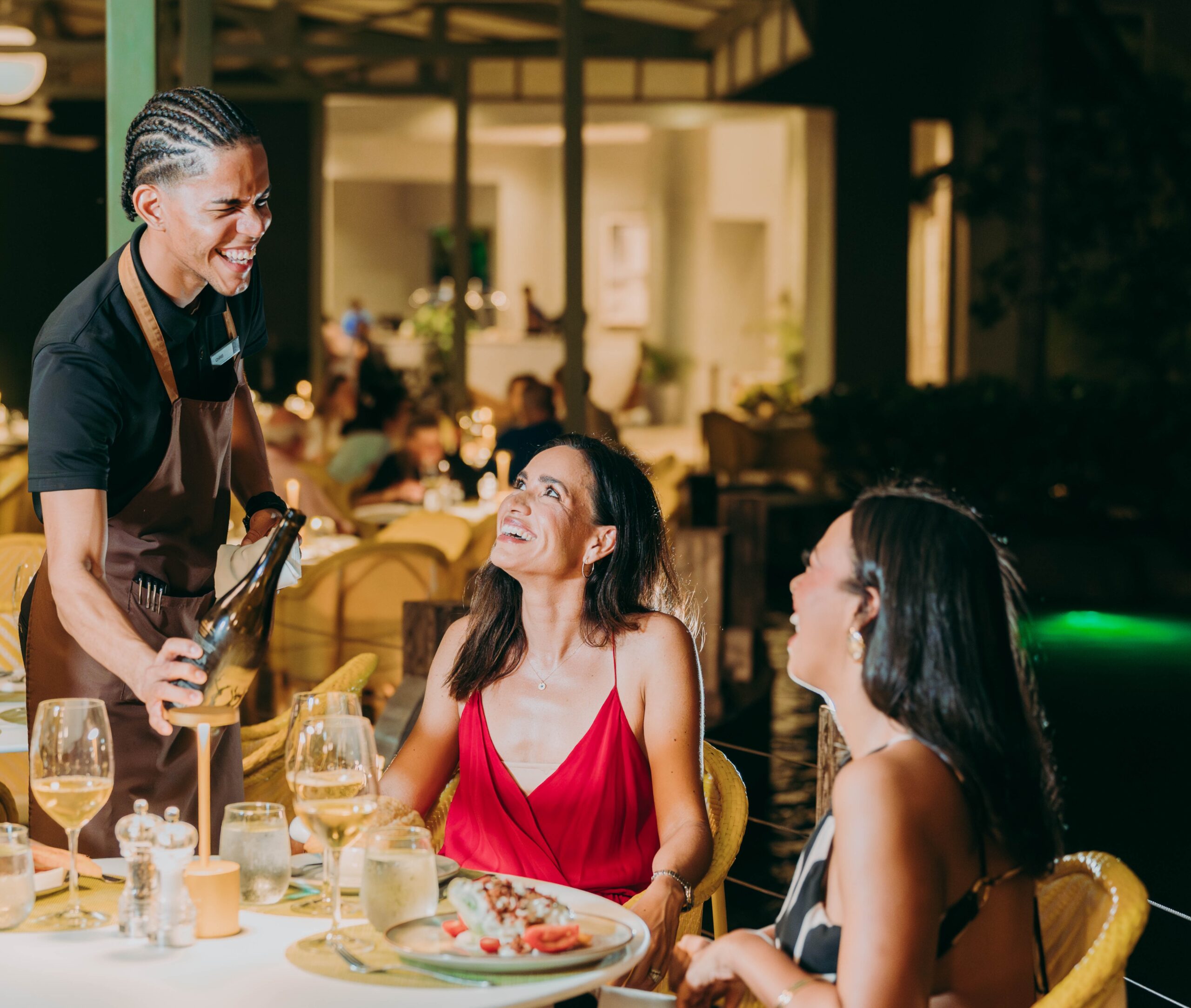 A smiling waiter pours wine for two women sitting at an outdoor restaurant table at night. The women are dressed elegantly and laughing, with plates of food and glasses in front of them. The atmosphere is warm and lively.