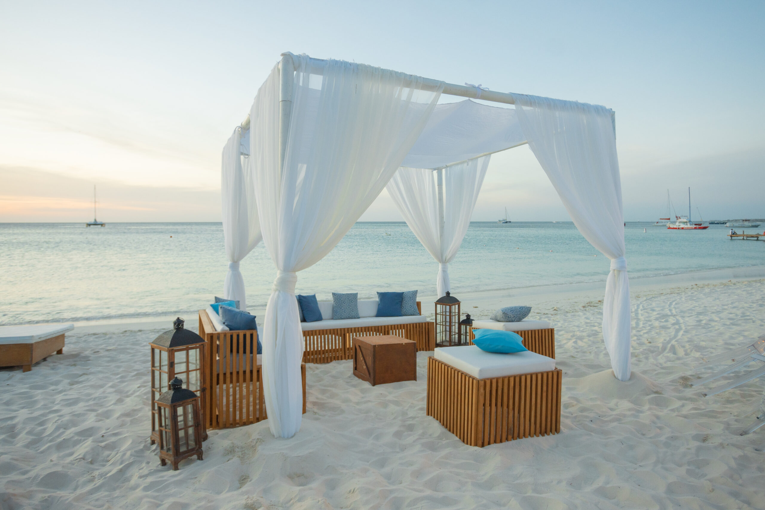 A white canopy with flowing curtains shelters wooden lounge furniture and blue pillows on a sandy beach, with lanterns nearby and sailboats on the calm sea in the background at sunset.