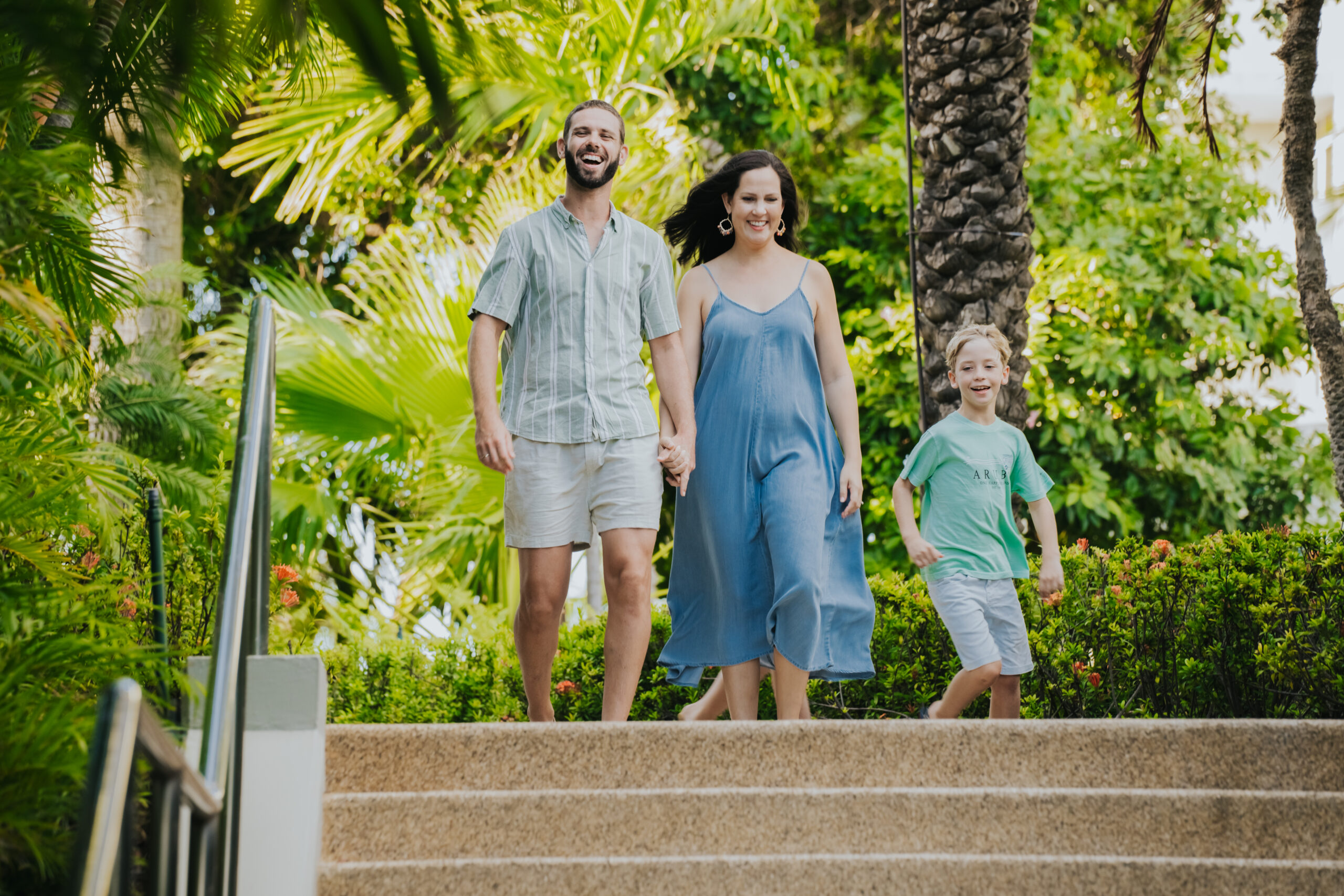 A smiling family of three walks down outdoor steps surrounded by lush green trees and plants. The adults hold hands while the child walks beside them, all dressed in casual summer clothing.