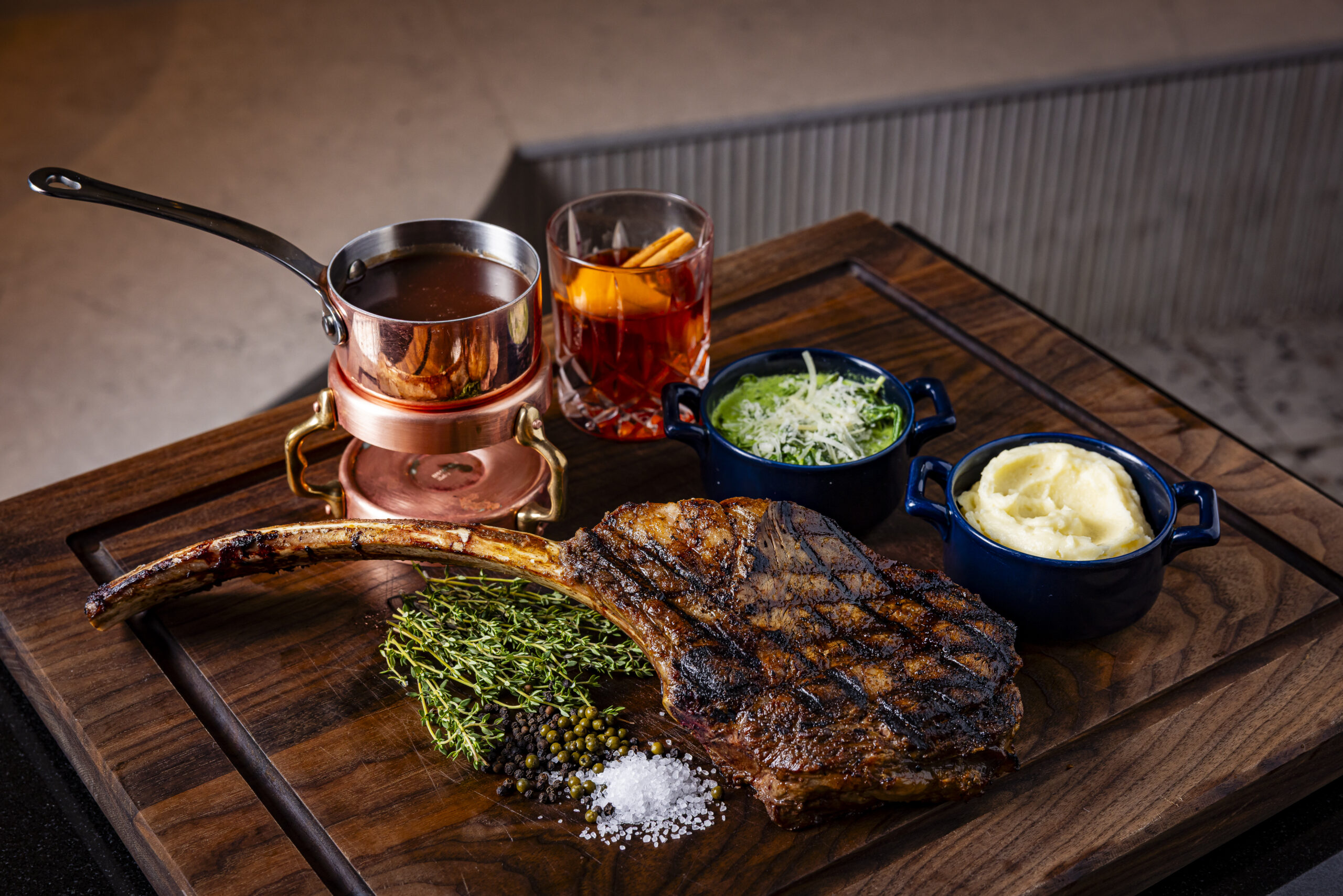 A grilled tomahawk steak on a wooden board with mashed potatoes, sautéed greens, a copper sauce pot, a glass of cocktail with orange slices, and garnishes of thyme, salt, and peppercorns.