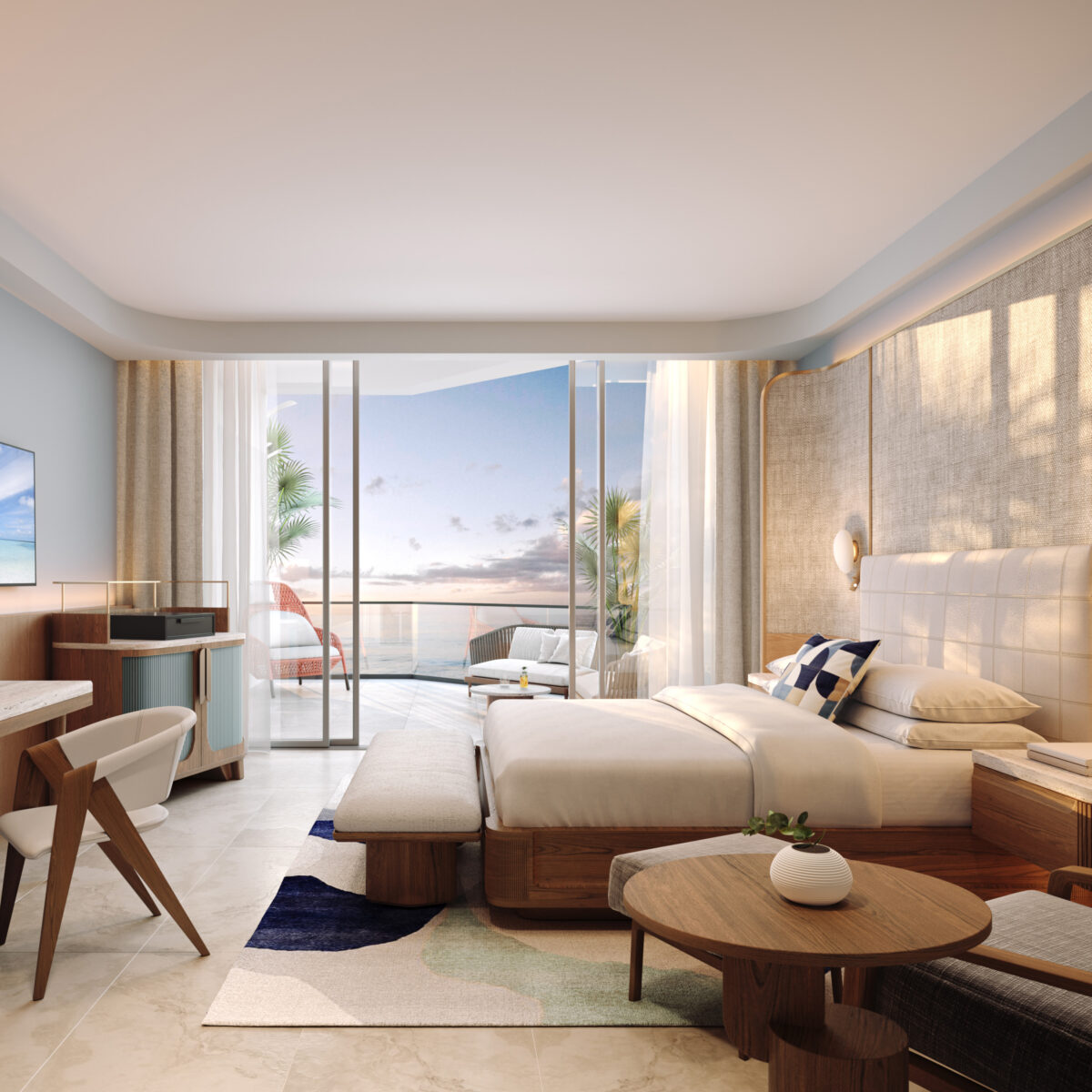 A modern hotel room with a large bed, desk, TV, and seating area. Floor-to-ceiling glass doors open to a balcony with ocean views at sunset. The decor features light colors and natural wood accents.