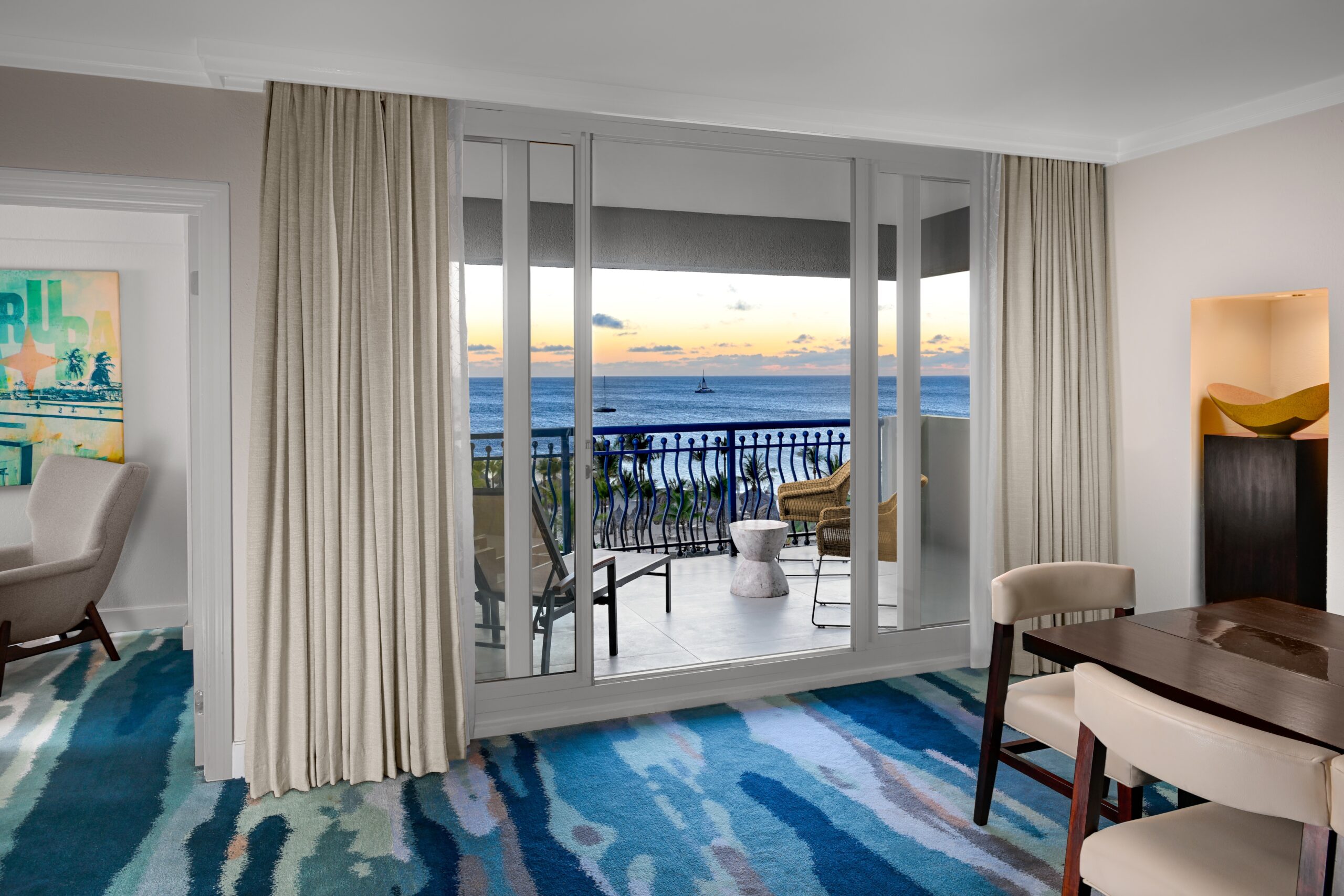 A modern hotel room with a dining table and chairs, patterned blue carpet, and a balcony with glass doors. The balcony overlooks the ocean at sunset, with a sailboat in the distance and outdoor seating visible.