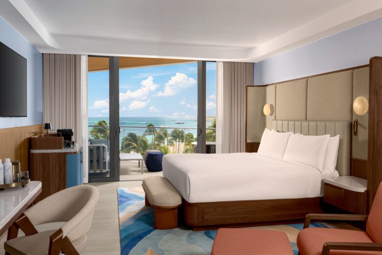 Modern hotel room with a large bed, seating area, TV, and coffee station. Floor-to-ceiling windows open to a balcony with a view of palm trees, blue sky, and the ocean.