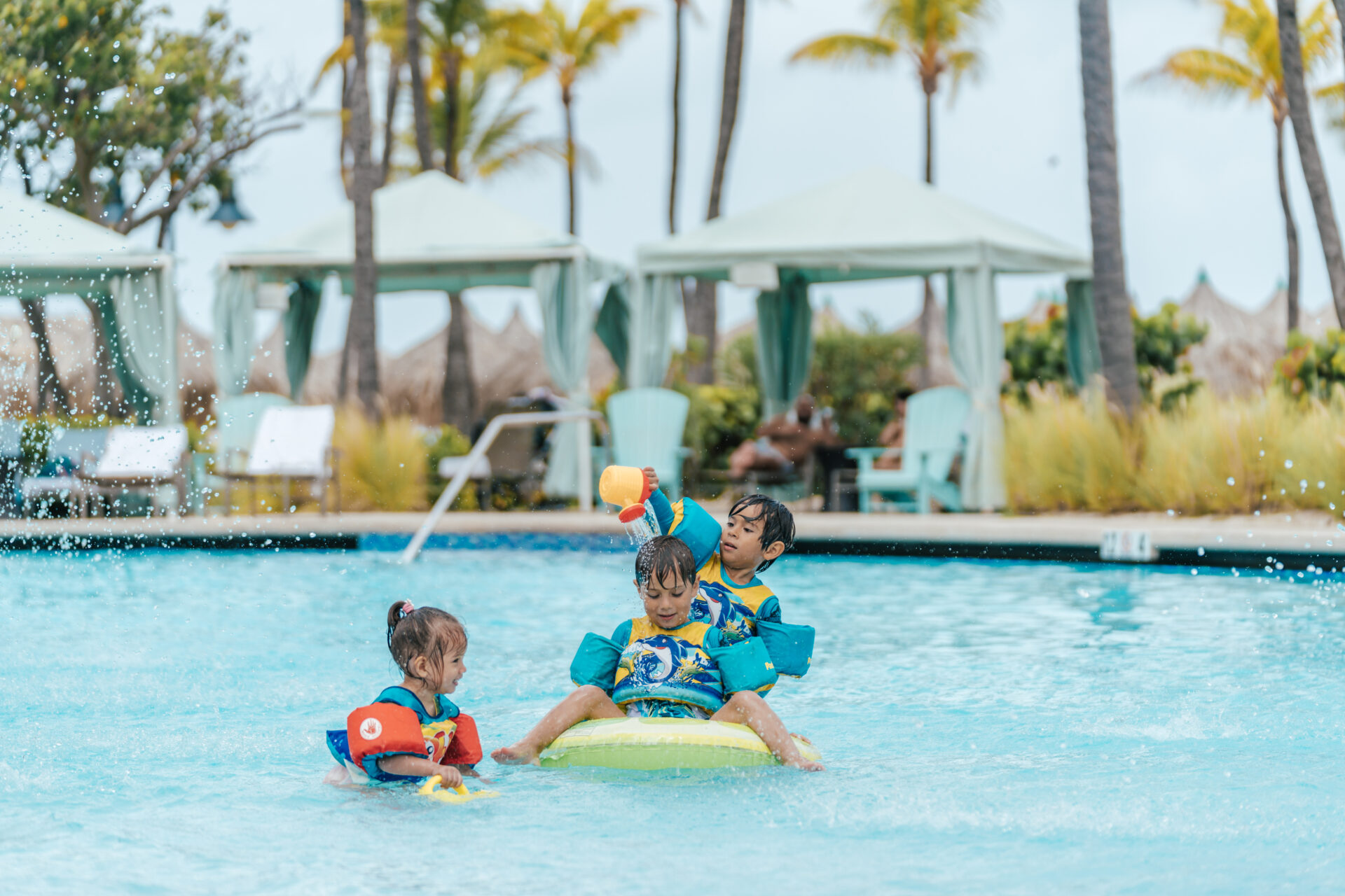 Three young children wearing floaties play together in a swimming pool, splashing water and laughing. Poolside cabanas, palm trees, and lounge chairs are visible in the background on a sunny day.