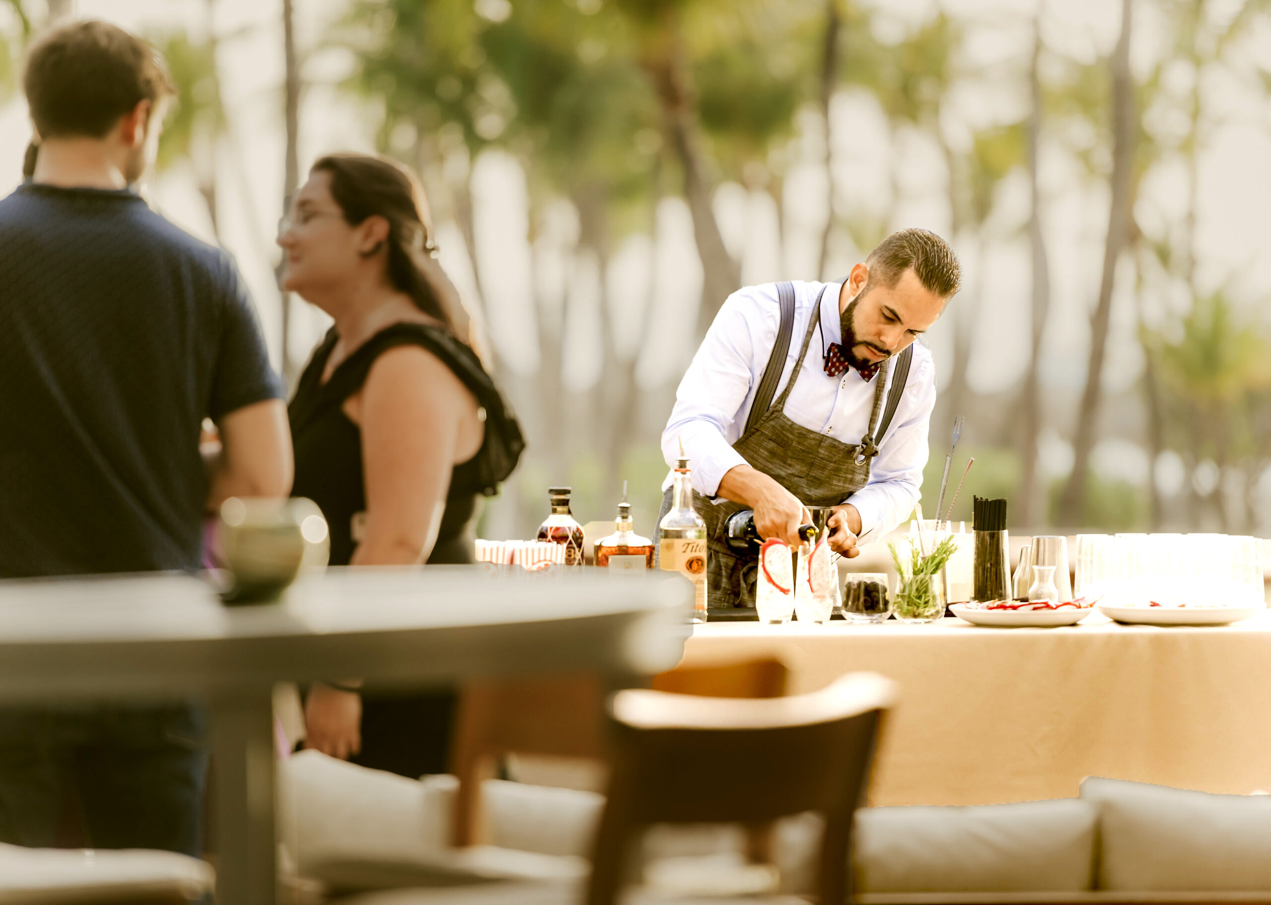 A bartender in a white shirt and bow tie prepares drinks at an outdoor bar while two people stand nearby; bottles and glasses are arranged on the table, with trees blurred in the background.