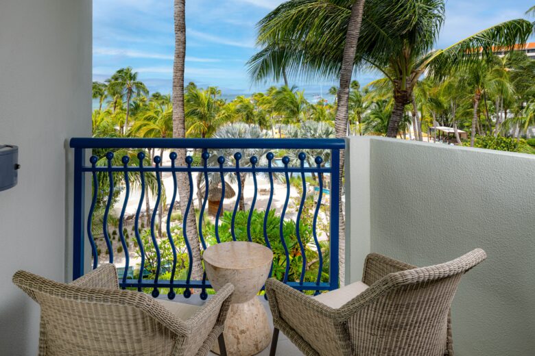 Two wicker chairs and a small round table are on a balcony with a blue railing, overlooking a lush tropical garden with palm trees and a view of the ocean in the background.