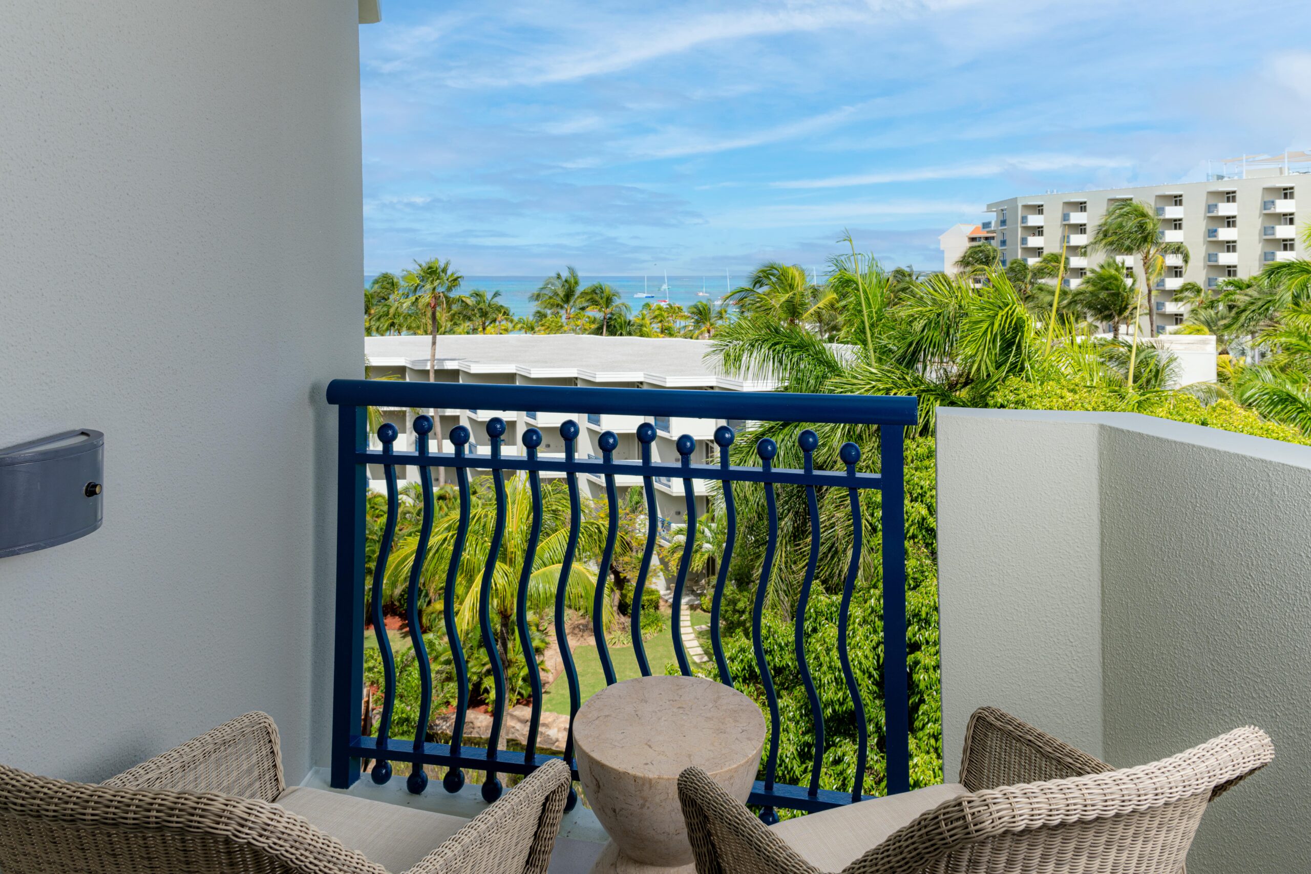 A small balcony with two wicker chairs and a round table overlooks lush greenery and palm trees, with modern buildings and the ocean visible in the background under a partly cloudy blue sky.
