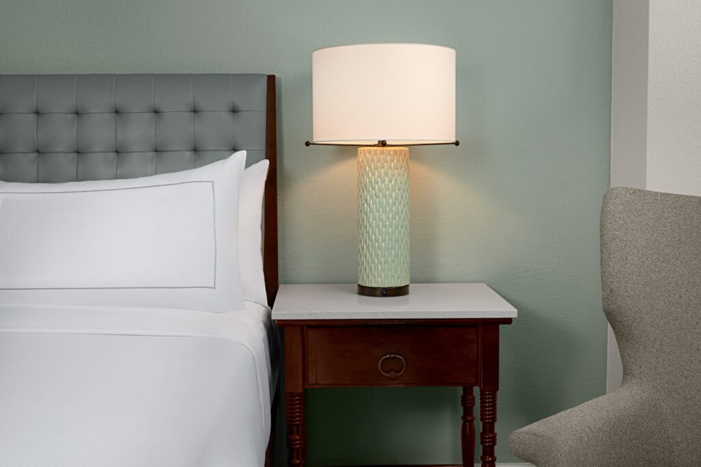 A neatly made bed with white linens sits beside a wooden nightstand holding a textured table lamp with a white shade. Part of a gray upholstered chair is visible on the right, against a soft green wall.