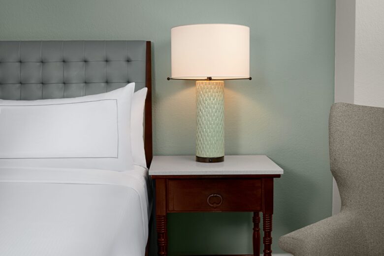 A neatly made bed with white linens sits beside a wooden nightstand holding a textured table lamp with a white shade. Part of a gray upholstered chair is visible on the right, against a soft green wall.