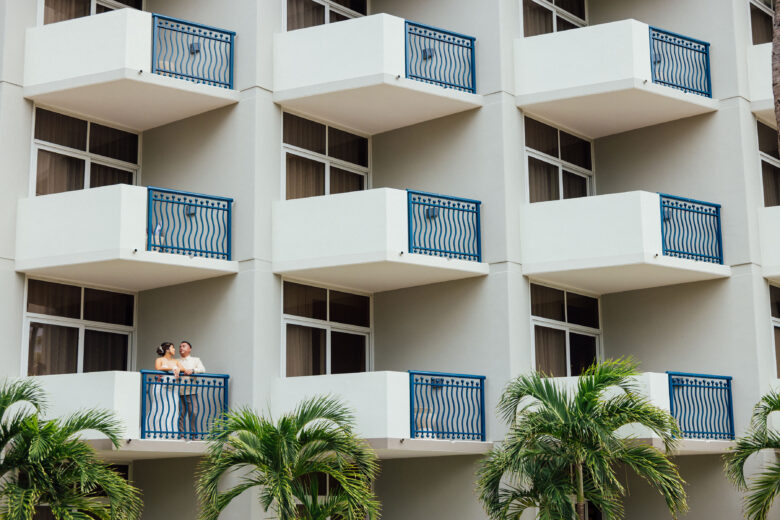 Two people stand together on a balcony of a modern building with multiple white balconies and blue railings; palm trees are visible in front of the building.