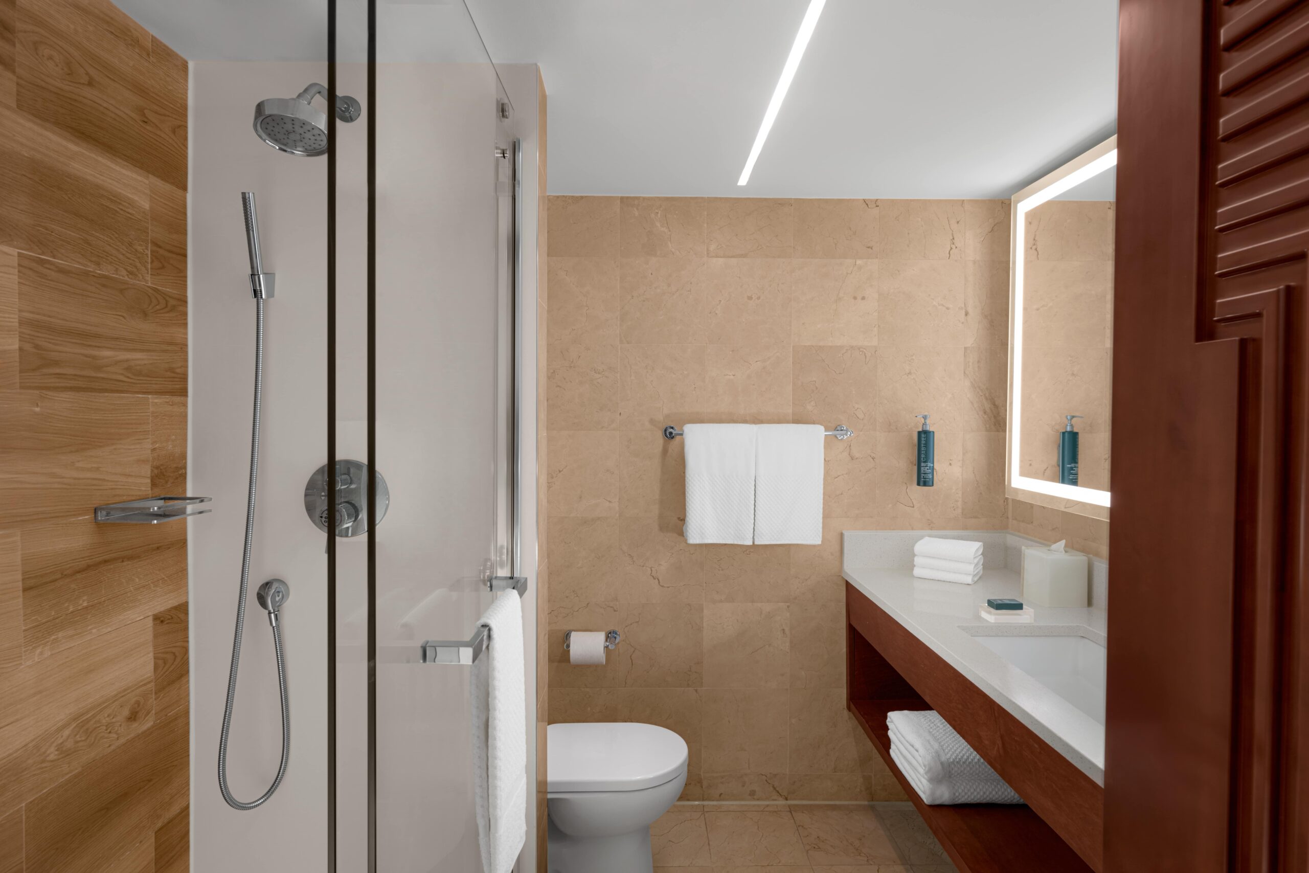 Modern bathroom with a glass-enclosed shower, wall-mounted showerhead, beige tile walls, white toilet, large mirror with lights, white countertop, towels, and toiletries neatly arranged.