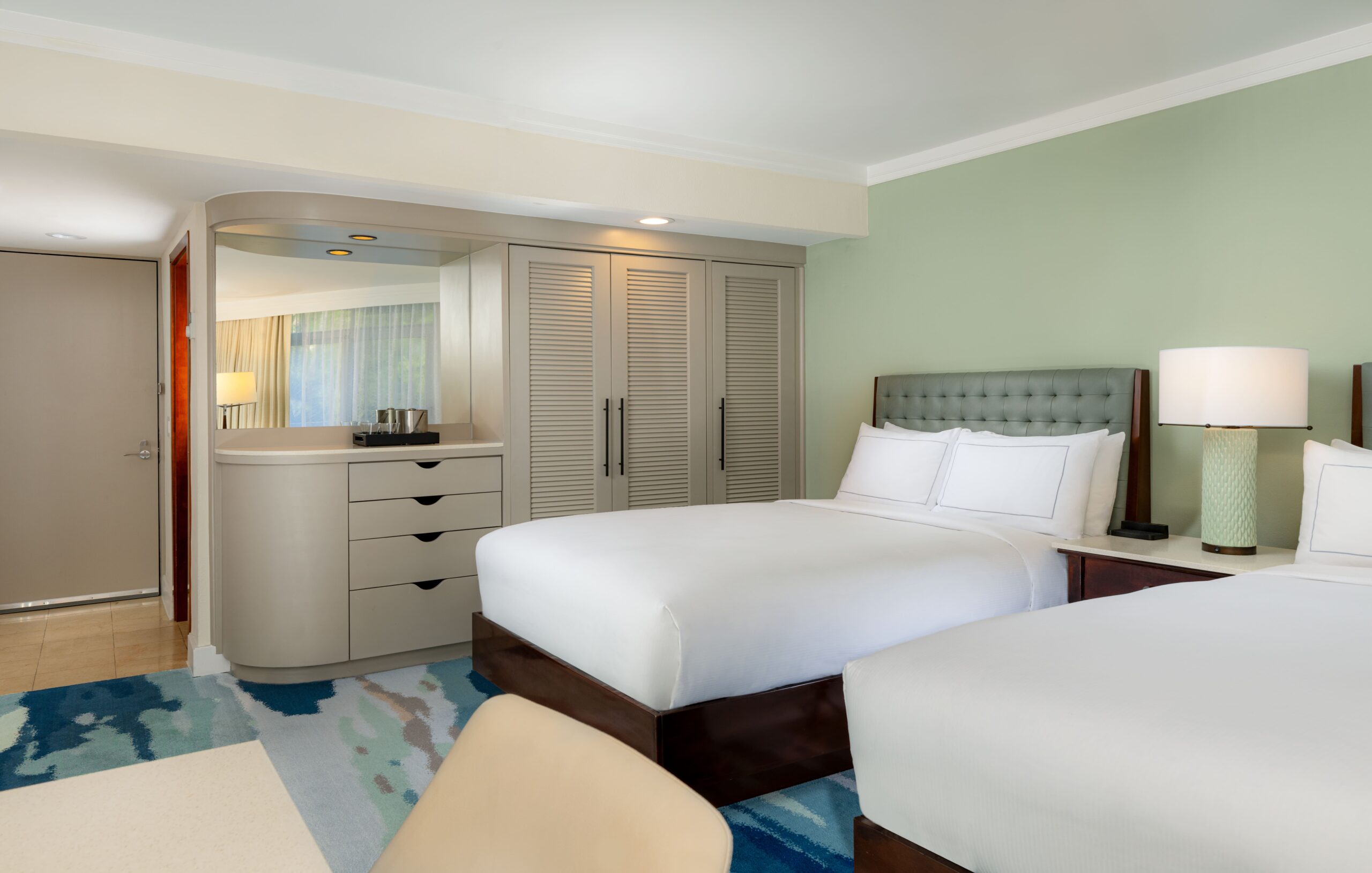 Modern hotel room with two double beds, white linens, tufted headboards, bedside lamps, a dresser with a large mirror, and a closet with louvered doors. The walls are light green and the floor has a blue patterned carpet.