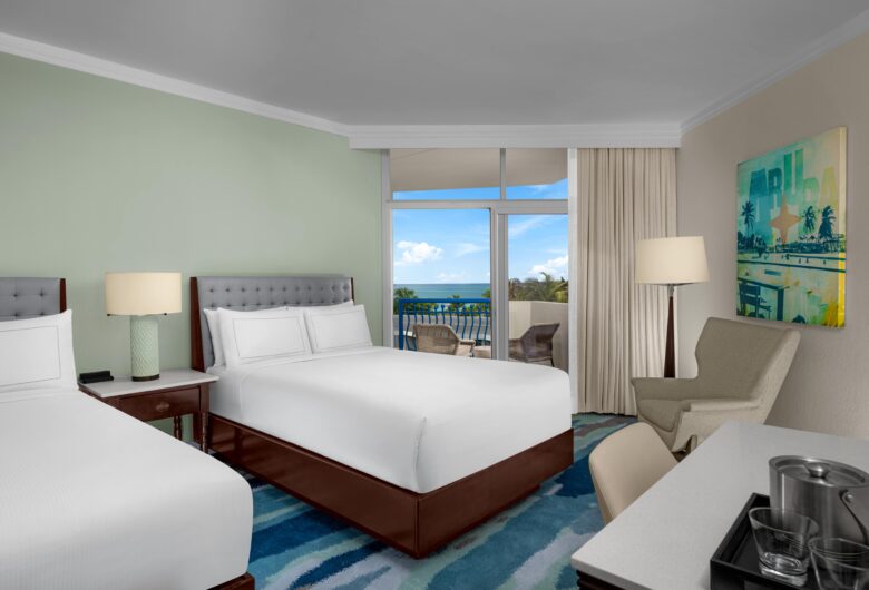 Hotel room with two double beds, bedside tables and lamps, an armchair, a desk with a tray and ice bucket, a large window with curtains, and a balcony with a view of the ocean and blue sky.