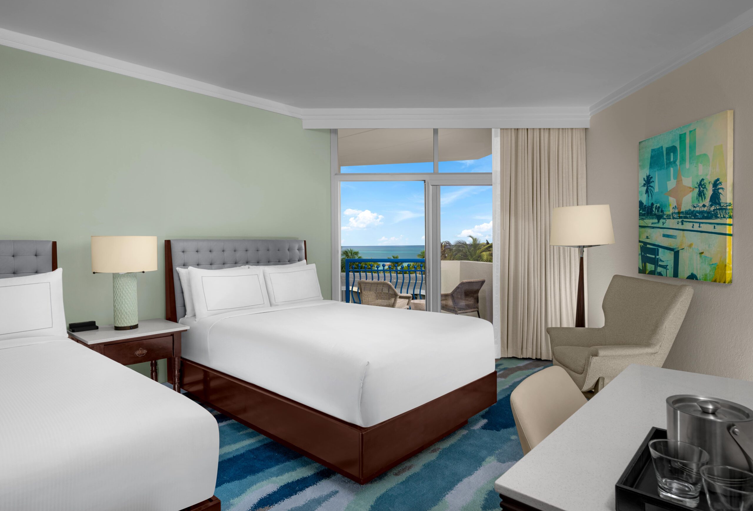 Hotel room with two double beds, bedside tables and lamps, an armchair, a desk with a tray and ice bucket, a large window with curtains, and a balcony with a view of the ocean and blue sky.