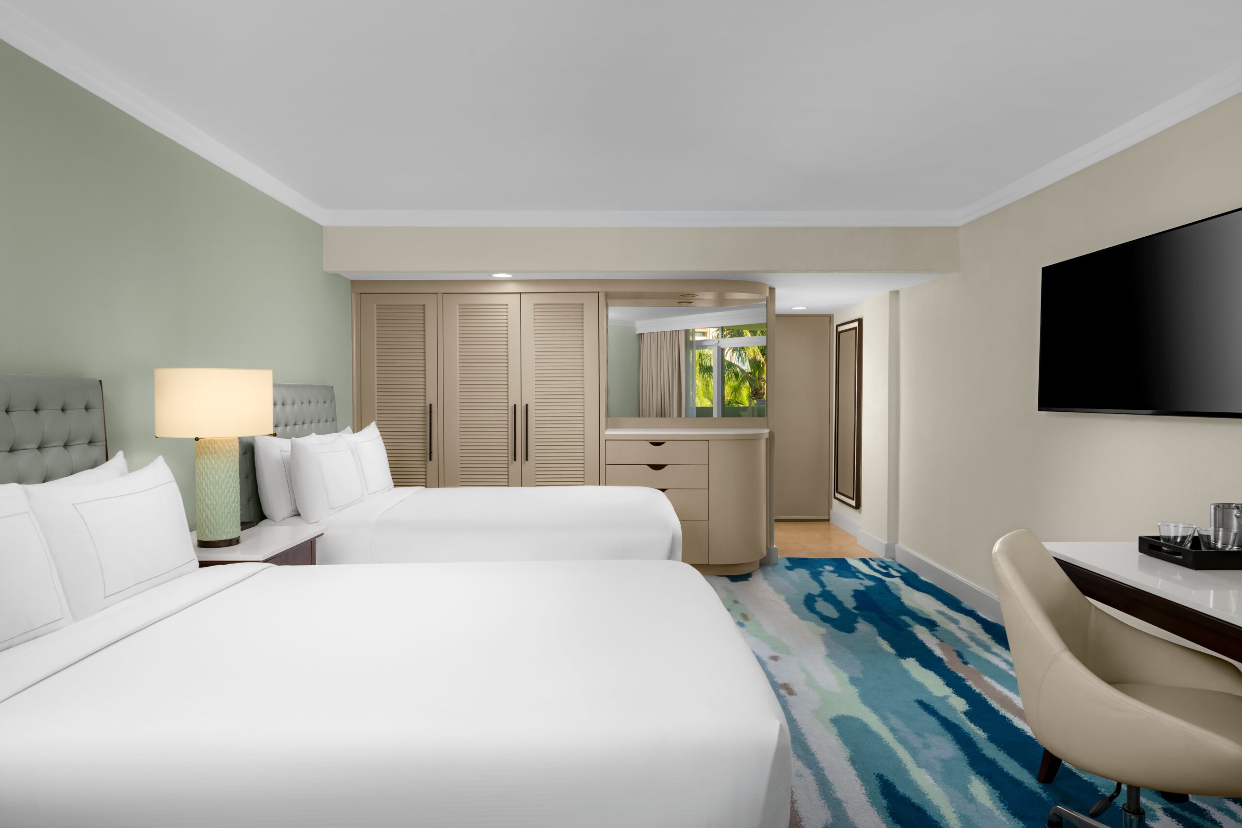 A hotel room with two neatly made double beds, a light green accent wall, a large flat-screen TV, a desk with a chair, a dresser, and a window showing greenery outside.