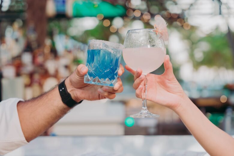 Two people clink glasses, one holding a blue cocktail in a short glass with a salted rim, the other holding a pink drink in a large wine glass, against a blurred outdoor bar background with bokeh lights.