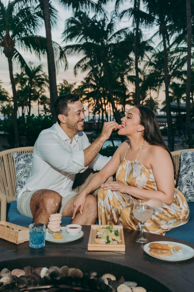 A man and woman sit outdoors by a table with food and drinks, surrounded by palm trees at sunset. The man is smiling and feeding the woman, who is also smiling, as they enjoy a relaxed, tropical setting together.