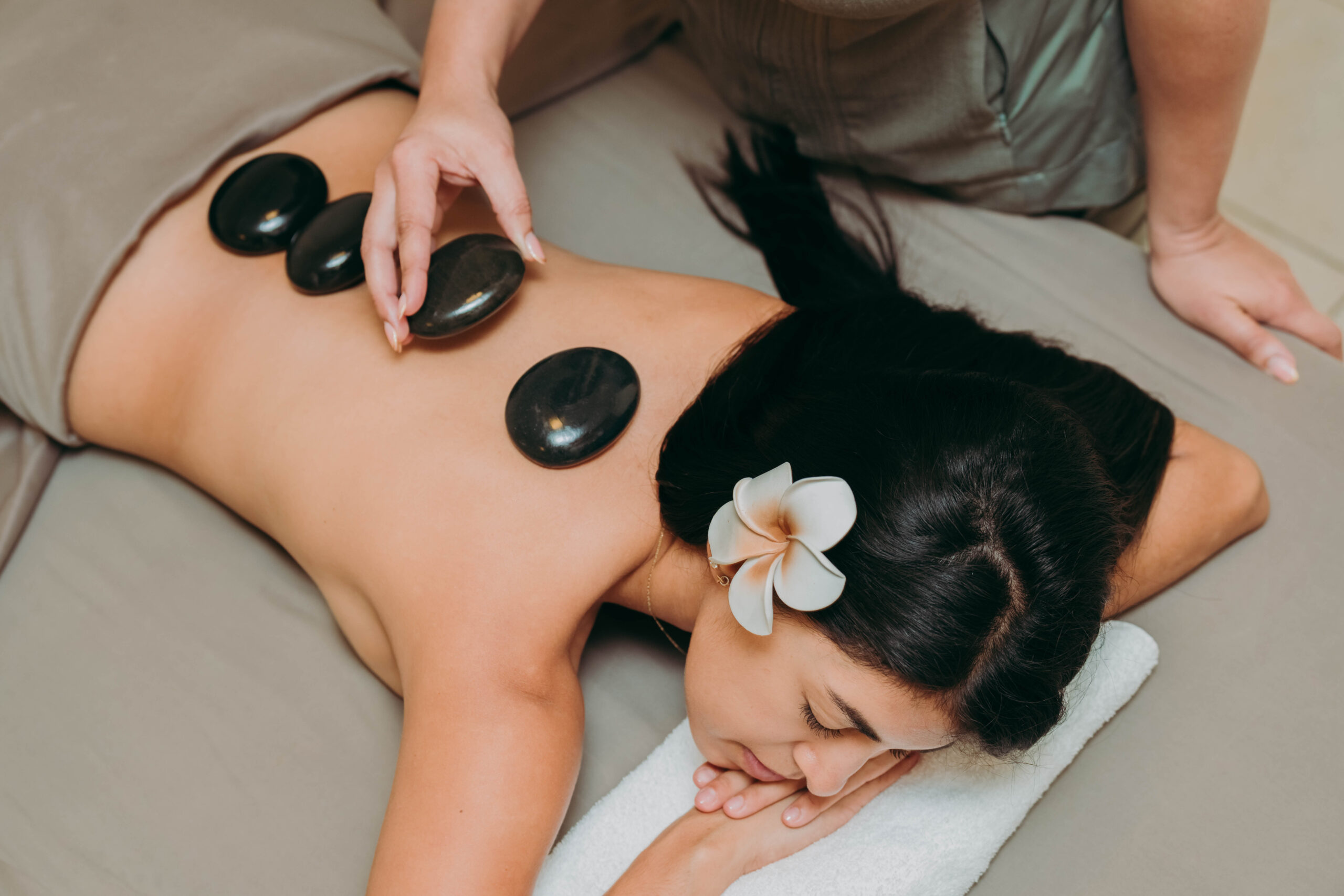 A woman with a white flower in her hair lies face down on a massage table while a therapist places smooth hot stones along her bare back during a spa treatment.
