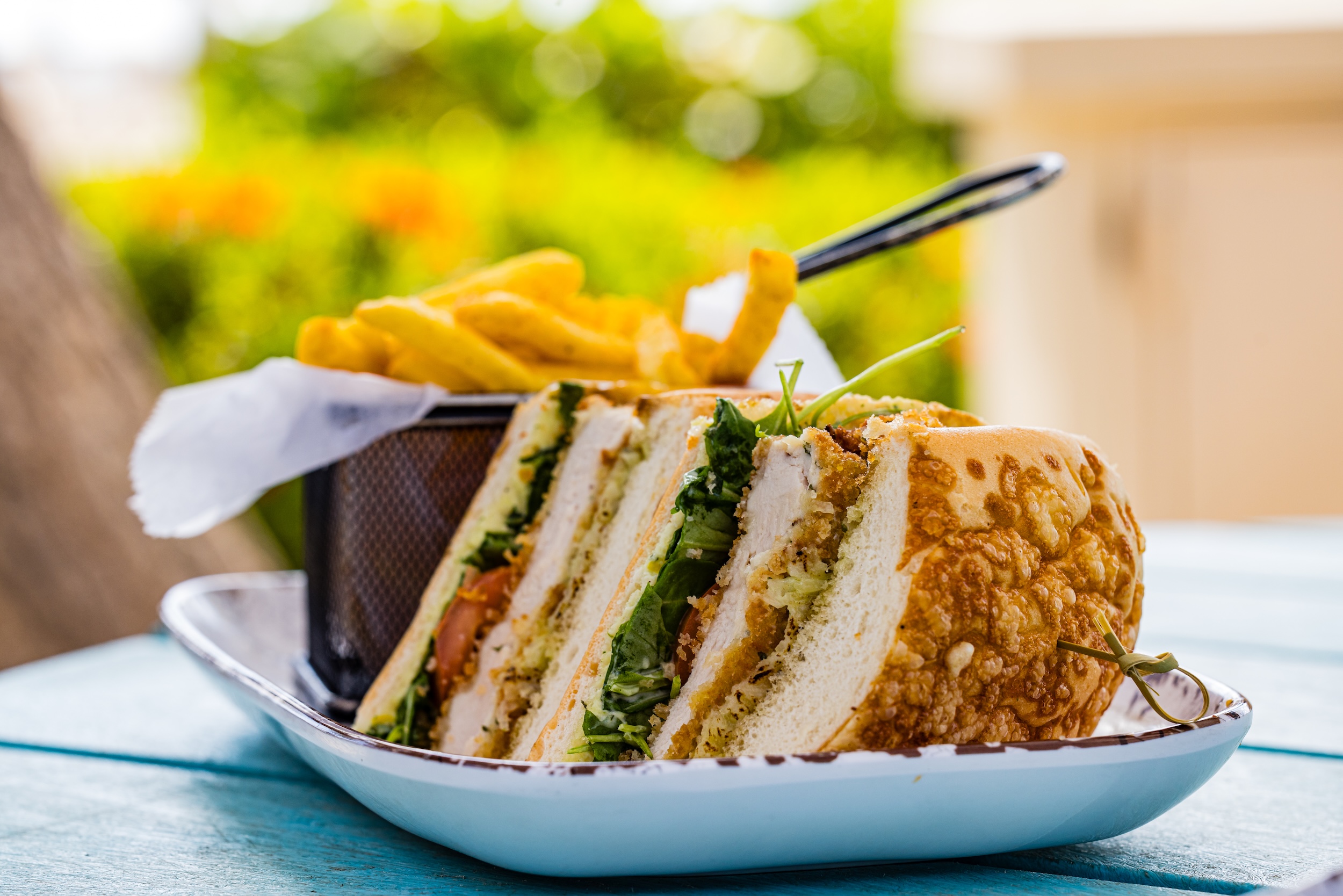 A close-up of a sandwich filled with lettuce, tomato, and chicken, cut into quarters and served on a white dish next to a small basket of French fries, with a blurred outdoor background.