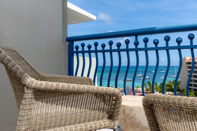 Two wicker chairs and a small table on a balcony with a blue railing, overlooking a turquoise ocean with boats and a coastline under a bright, sunny sky.