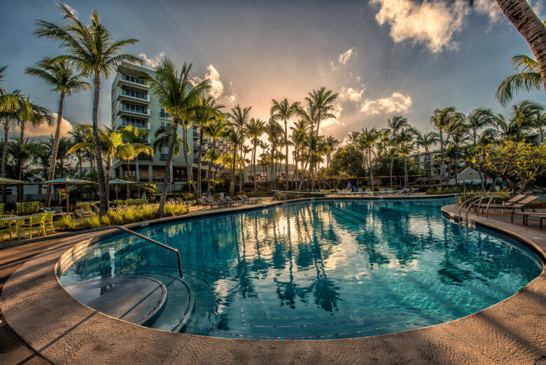 A resort swimming pool surrounded by palm trees and lounge chairs at sunset, with tall buildings in the background and a clear blue sky reflecting in the water.