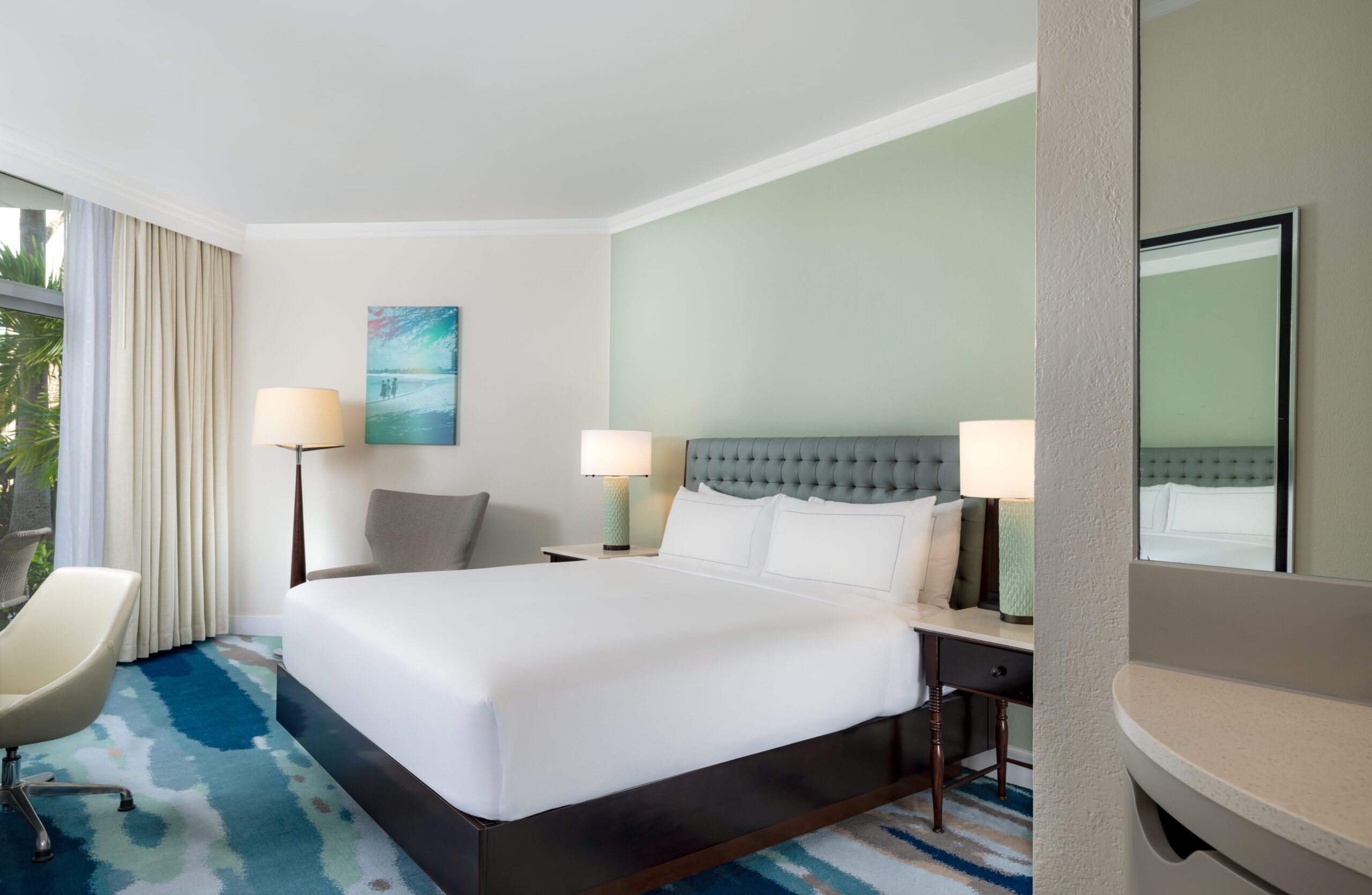 A modern hotel room with a large bed, white bedding, two bedside tables with lamps, an armchair, a desk chair, a large window with curtains, and abstract art on a soft green accent wall.