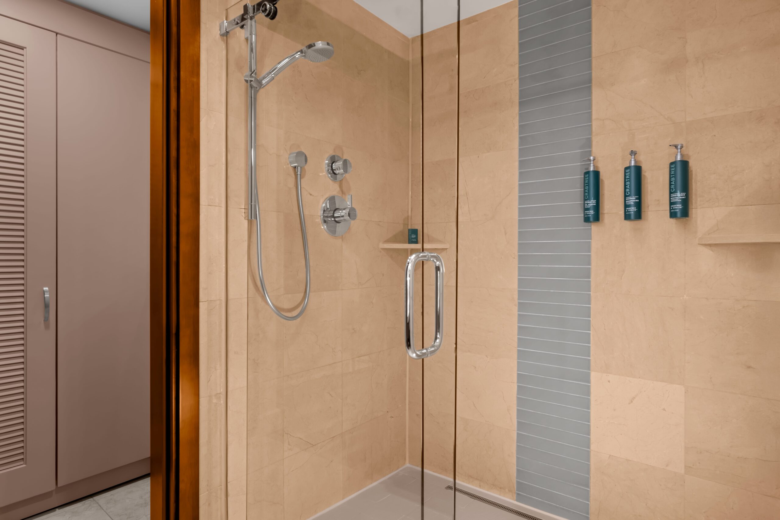 A modern shower with beige tiles, a glass door, a silver showerhead, control knobs, built-in shelves, and three green bottles mounted on the wall. A pink closet is visible outside the shower area.
