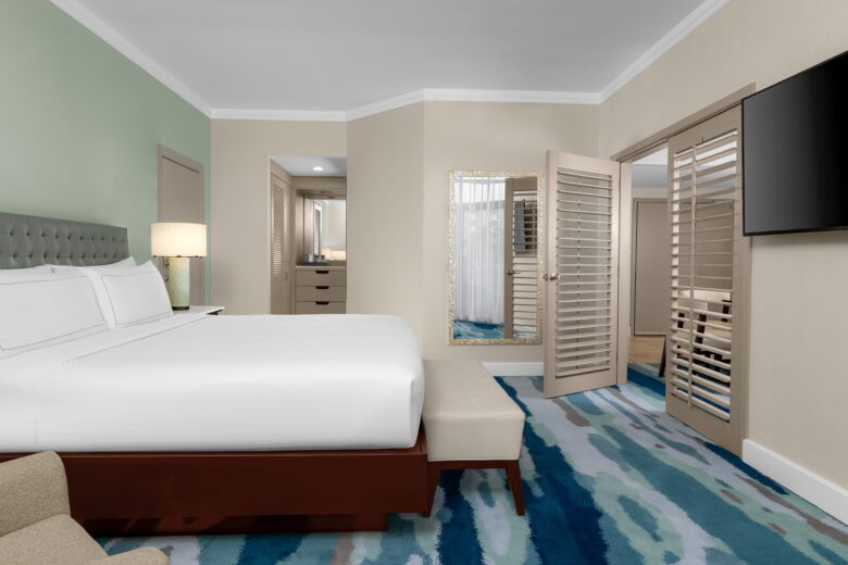 A modern hotel room with a large bed, patterned blue carpet, a bench at the foot of the bed, a flat-screen TV on the wall, a lamp, a mirror, and open shuttered doors leading to another room.