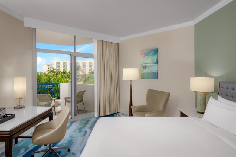 A bright hotel room with a large bed, work desk, chairs, lamps, and a balcony with outdoor seating. The balcony overlooks palm trees and another building. Walls are light, and a colorful painting hangs above a chair.