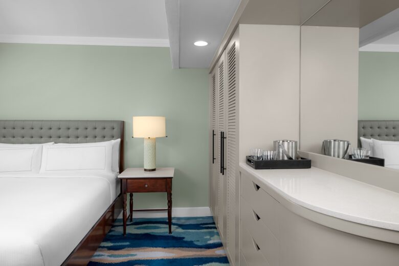 A modern hotel room with a neatly made bed, a bedside table with a lit lamp, a closet with louvered doors, and a countertop with a tray holding glasses and an ice bucket. A blue patterned rug is on the floor.