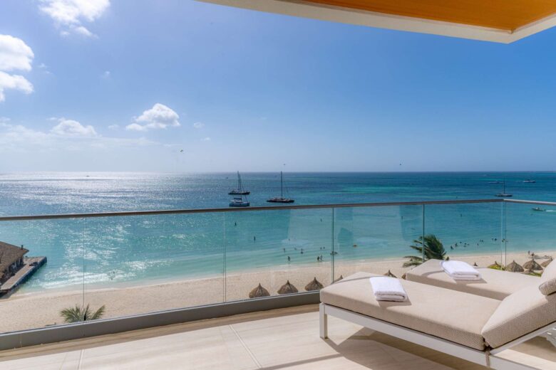 Two lounge chairs with towels overlook a sandy beach and turquoise ocean. Sailboats float on the water under a blue sky, seen from a balcony with glass railing.