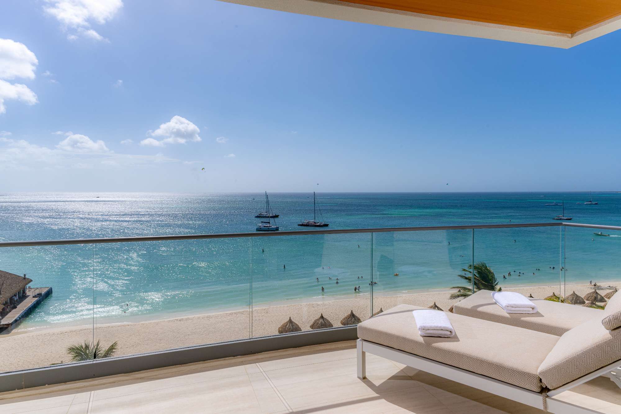 Two lounge chairs with towels overlook a sandy beach and turquoise ocean. Sailboats float on the water under a blue sky, seen from a balcony with glass railing.