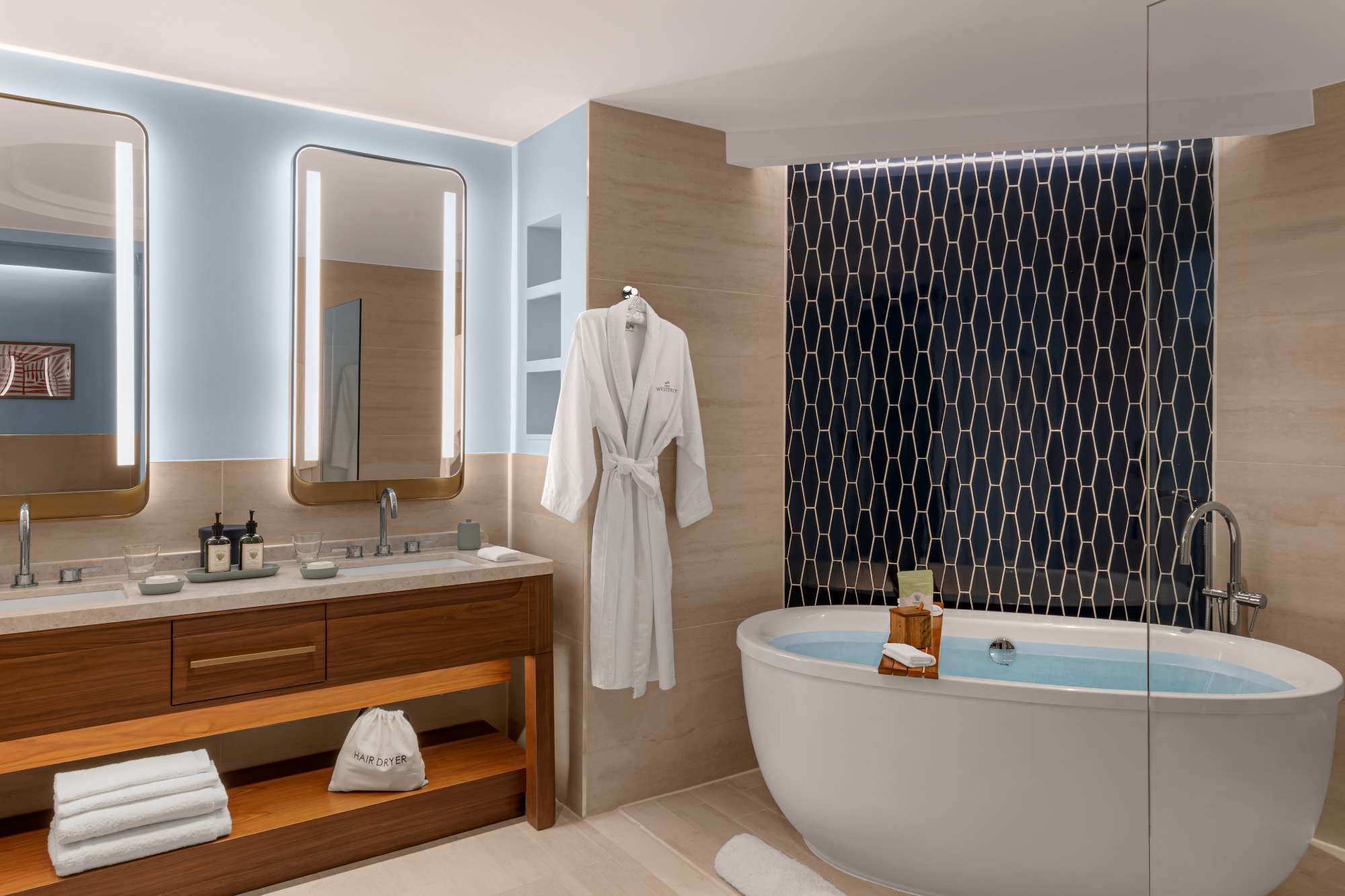 Modern bathroom with a freestanding tub, dark tiled accent wall, wooden vanity with double sinks, large mirrors, towels, bathrobe on a hook, and soft lighting.